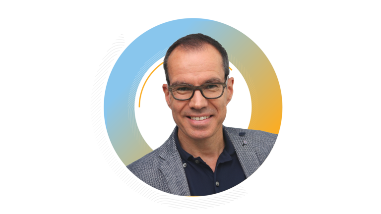 Porträt von Boris Felgendreher lächelnd, mit Brille und grauem Sakko, vor einem kreisförmigen blau-orangenen Hintergrund.